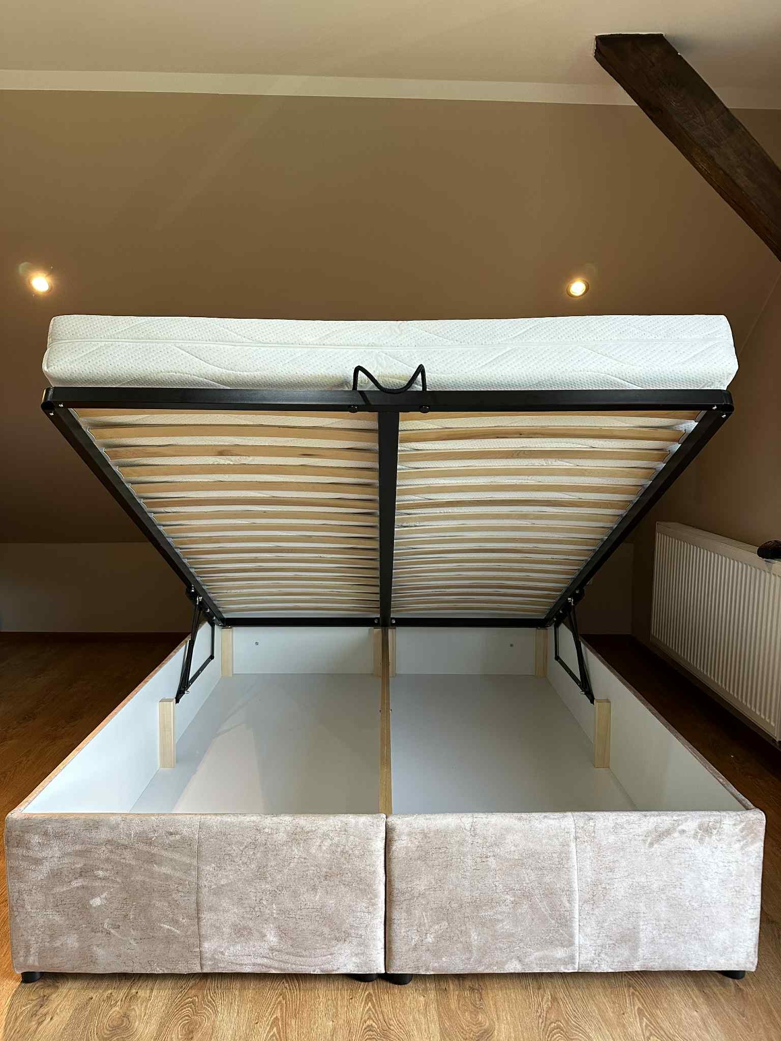 Otwarta rama łóżka tapicerowanego z pojemnikiem, widoczne drewniane listwy i mechanizm podnoszenia. Beżowa tapicerka i drewniana podłoga w tle.