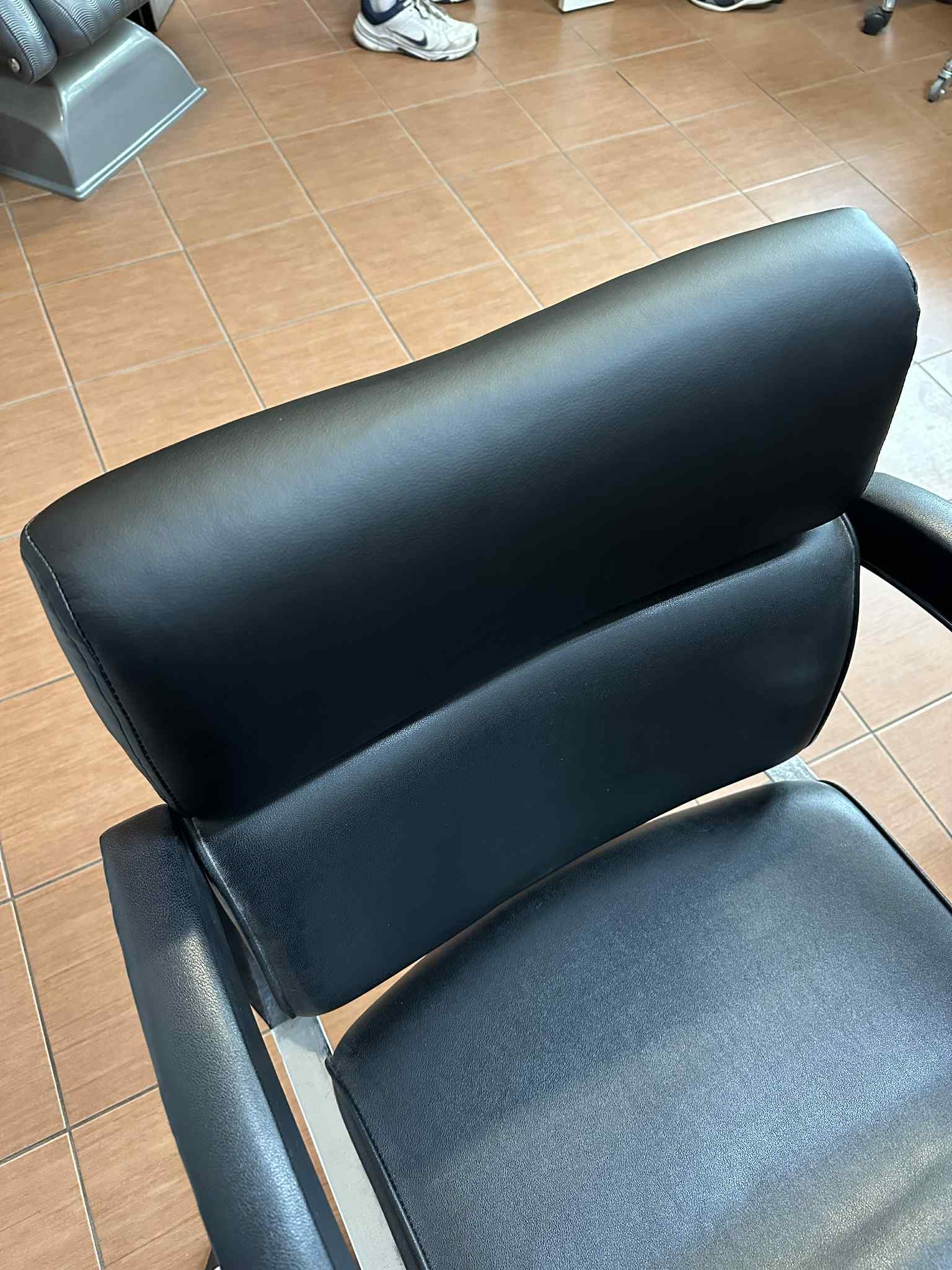 Czarne, skórzane krzesło fryzjerskie z widocznym zagłówkiem i podłokietnikami, na tle jasnobrązowych płytek podłogowych. Widok z góry na oparcie krzesła.