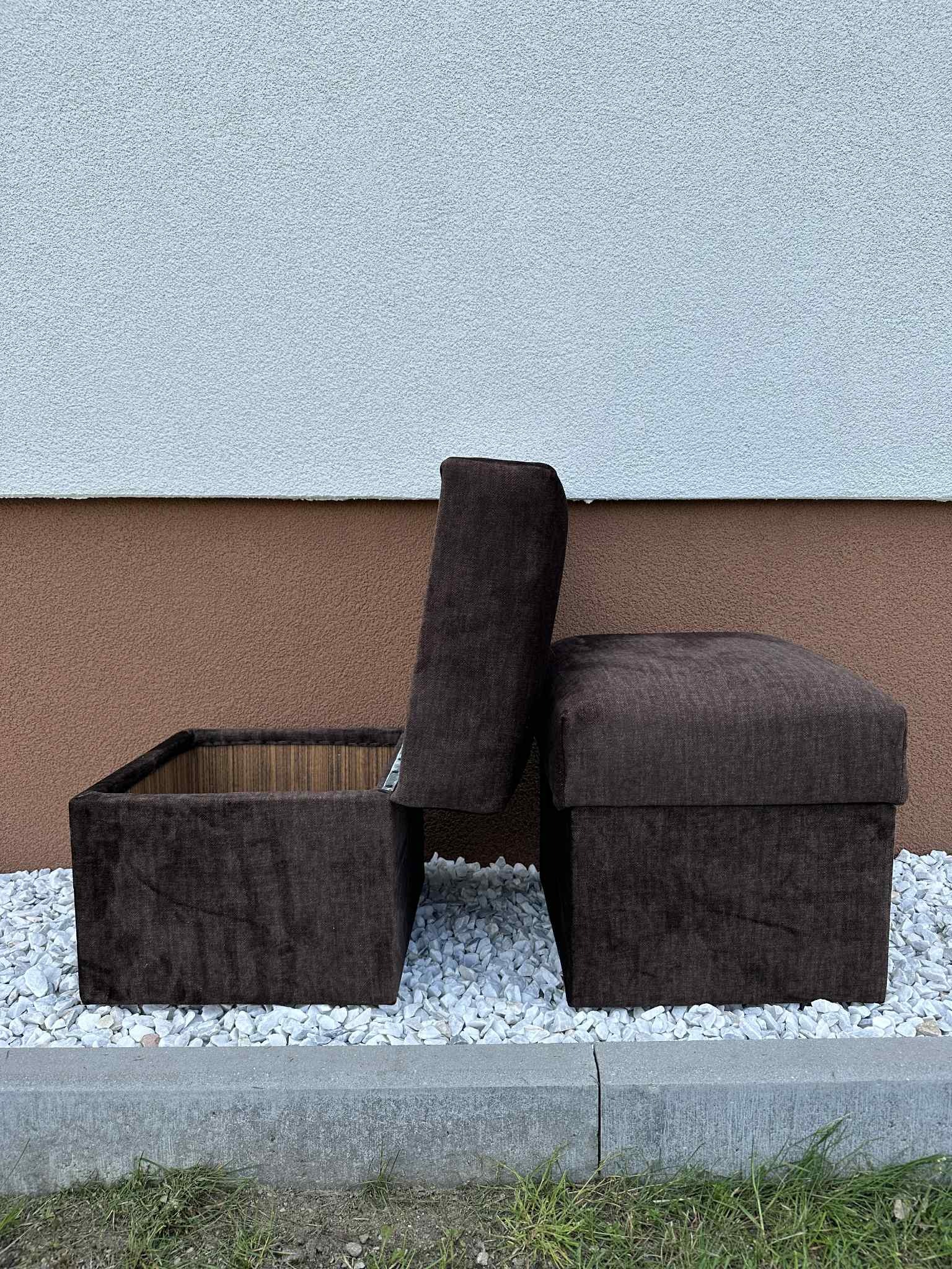 Brązowy, tapicerowany puf z otwieranym schowkiem, stojący na białym żwirze przed budynkiem. Widoczny otwarty schowek z drewnianym wykończeniem.