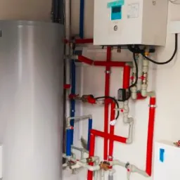 Biała pompa ciepła Panasonic zamontowana na ścianie, z widocznymi czerwonymi i niebieskimi rurami instalacji grzewczej oraz srebrnym zbiornikiem buforowym obok.