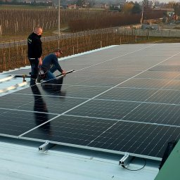 SOLMIL SPÓŁKA Z OGRANICZONĄ ODPOWIEDZIALNOŚCIĄ - Montaż paneli słonecznych na dachu budynku, dwóch instalatorów pracuje, jeden w masce, w tle widok na cmentarz i winnicę.