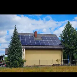 Dom z panelami fotowoltaicznymi na dachu, drabina oparta o ścianę, widok z poziomu gruntu, częściowo zasłonięty przez drzewa iglaste.