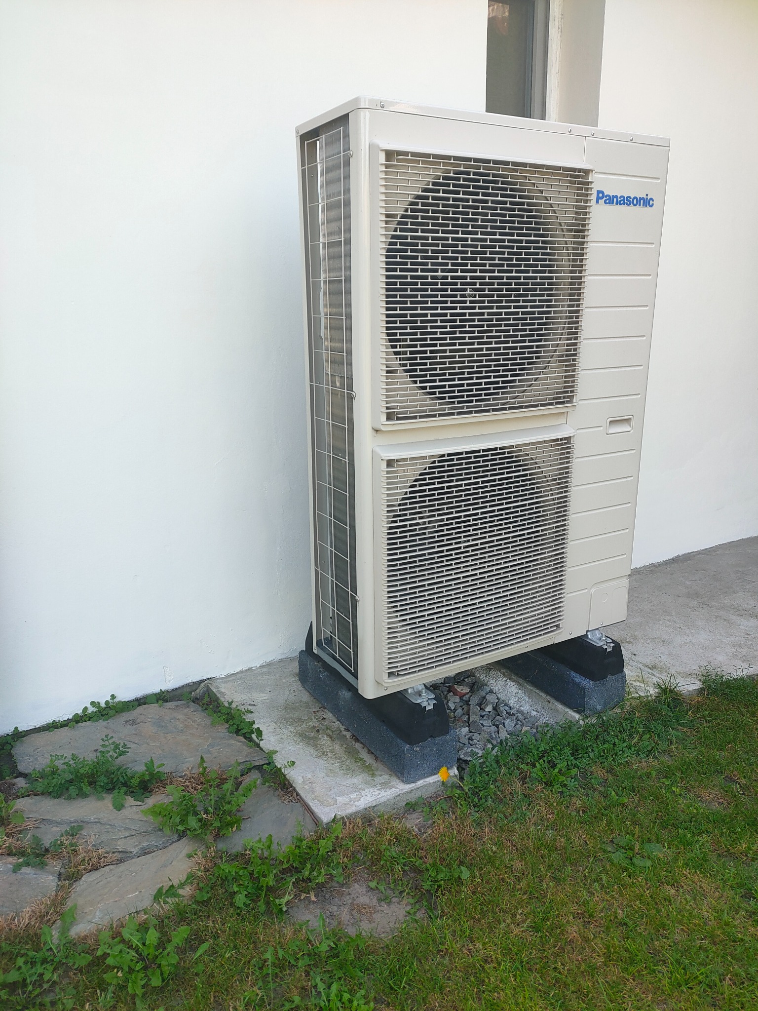Dwuczęściowa jednostka zewnętrzna klimatyzacji Panasonic, zamontowana na betonowej podstawie z gumowymi podkładkami, ustawiona na trawniku obok białej ściany budynku.