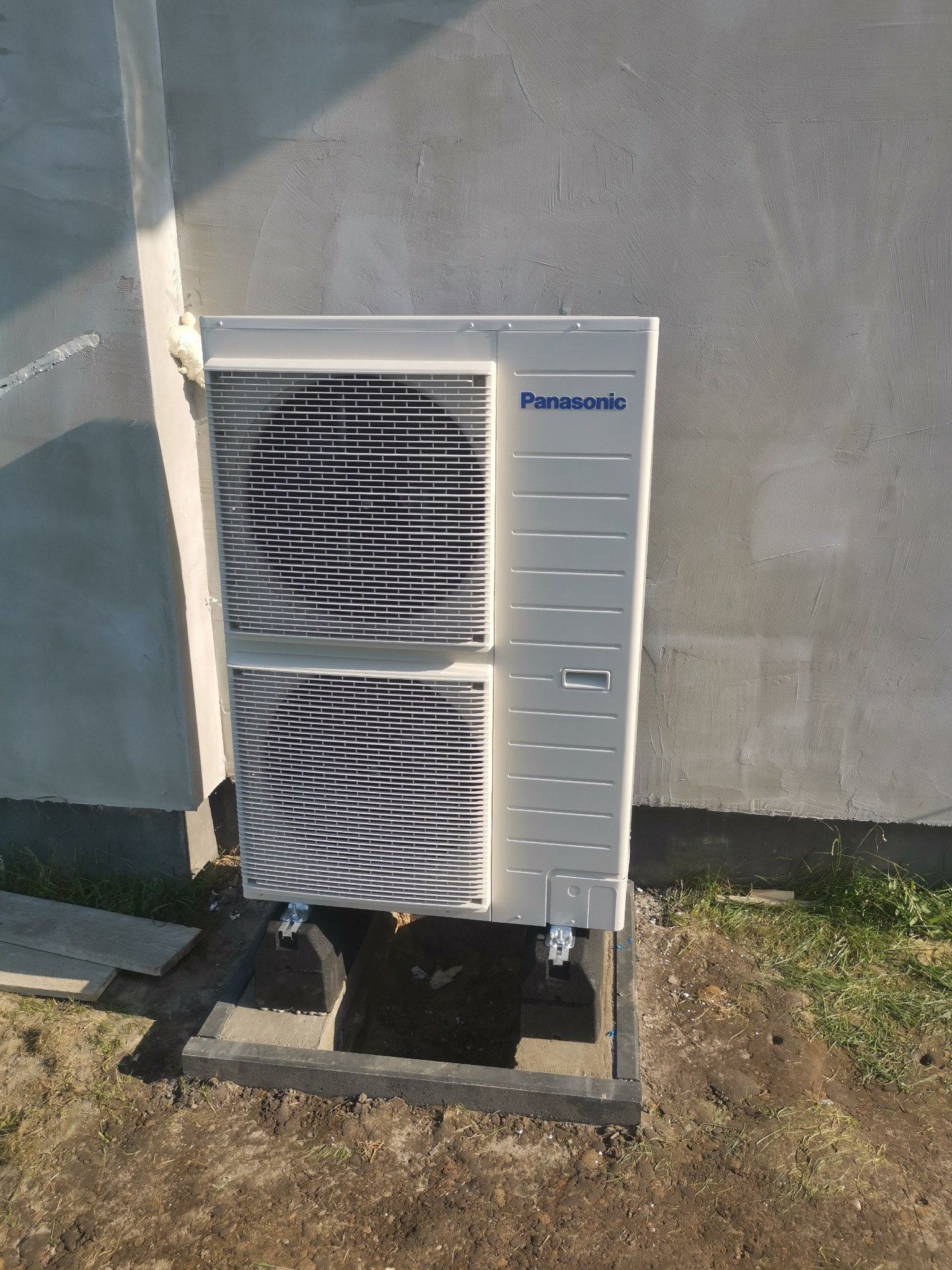 Biała jednostka zewnętrzna klimatyzacji marki Panasonic zamontowana przy szarej ścianie budynku, widoczne pianka montażowa i elementy instalacji w gruncie.