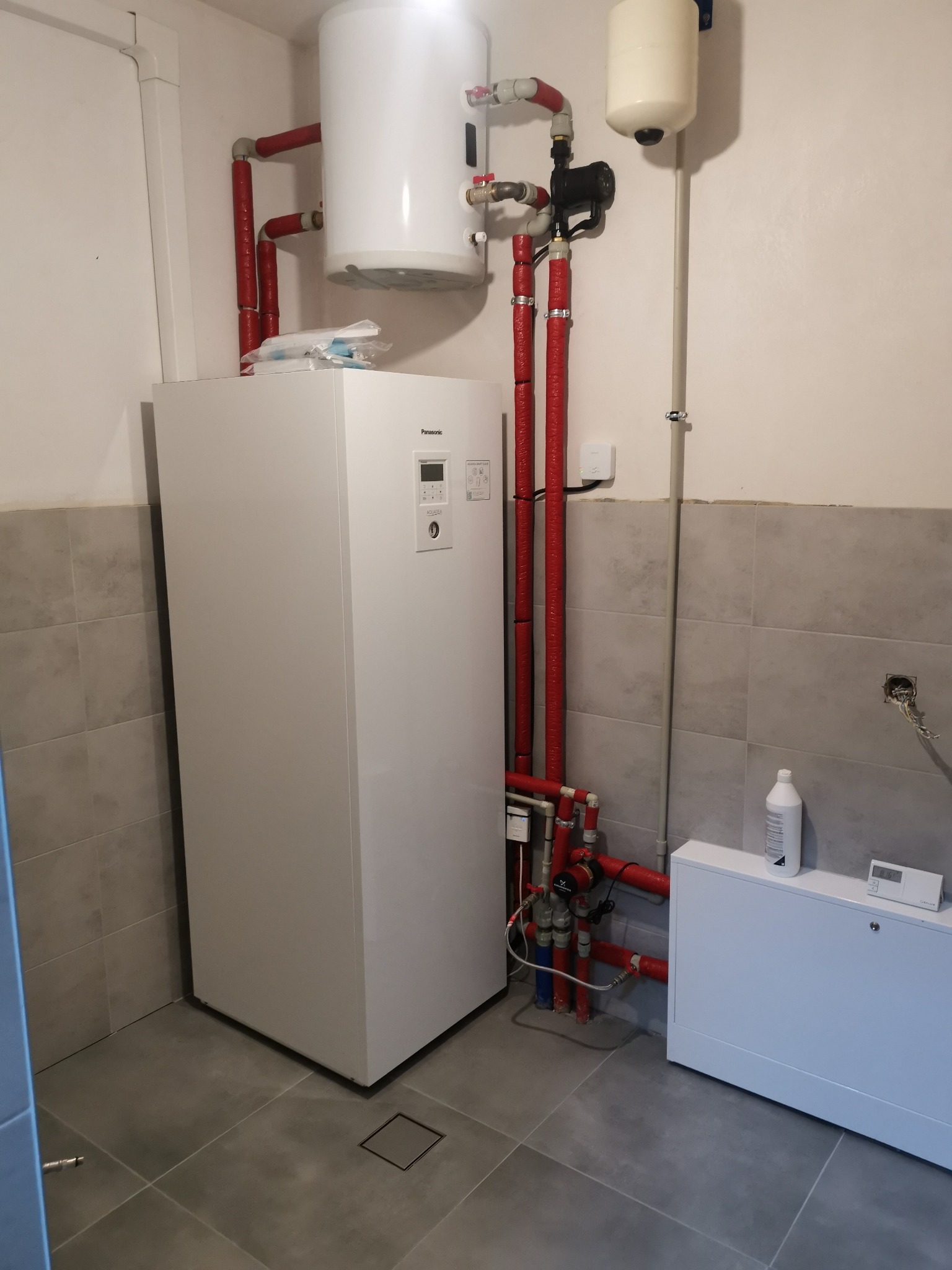 Biała pompa ciepła Panasonic Aquarea z czerwonymi rurami izolowanymi termicznie, zbiornik wyrównawczy i instalacja wodna w pomieszczeniu z szarą glazurą.