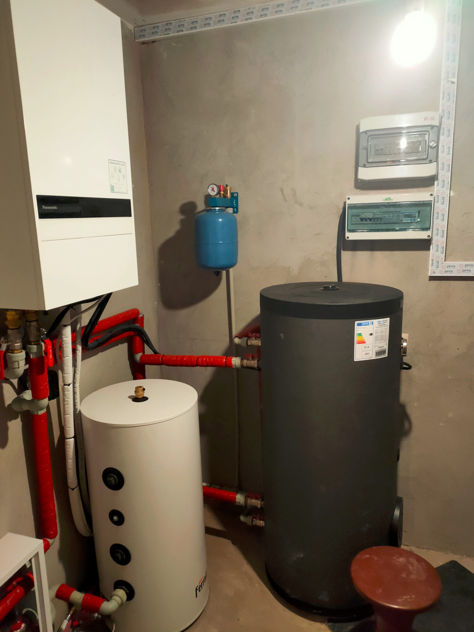 Wnętrze kotłowni z zamontowanym białym zasobnikiem ciepłej wody użytkowej marki Ferro, szarym zbiornikiem buforowym, białą pompą ciepła Panasonic oraz instalacją hydrauliczną z rur w izolacji,...