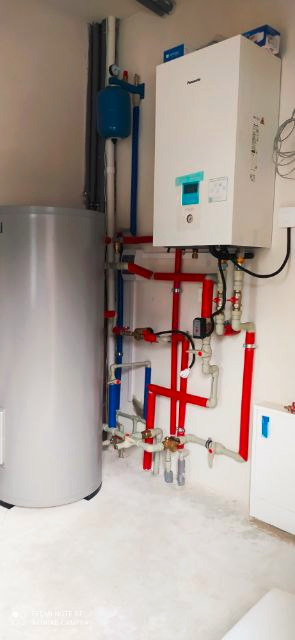 Biała pompa ciepła Panasonic zamontowana na ścianie, z widocznymi czerwonymi i niebieskimi rurami instalacji grzewczej oraz srebrnym zbiornikiem buforowym obok.