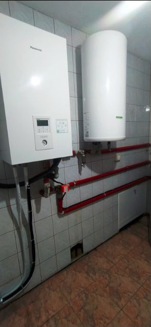 Biały piec centralnego ogrzewania Panasonic i pionowy zasobnik ciepłej wody, zamontowane na kafelkowanej ścianie, z widocznymi czerwonymi rurami instalacji grzewczej.