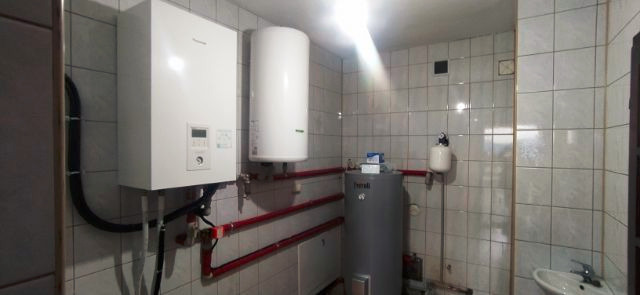 Wnętrze kotłowni z dwoma białymi kotłami grzewczymi marki Panasonic, szarym zasobnikiem ciepłej wody Ferroli, instalacją z rur w kolorze czerwonym i umywalką w rogu. Ściany wyłożone białymi...