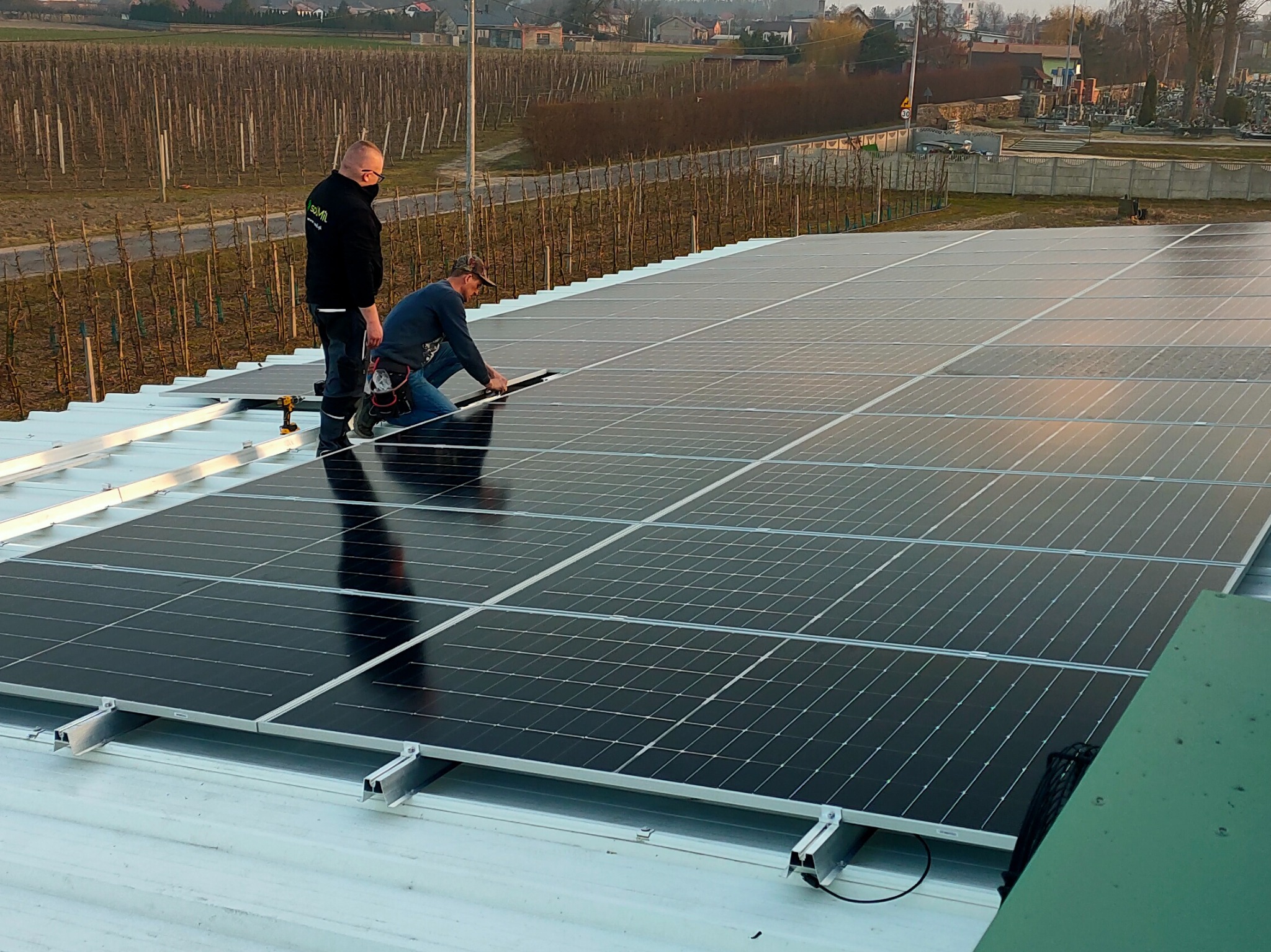 Montaż paneli słonecznych na dachu budynku, dwóch instalatorów pracuje, jeden w masce, w tle widok na cmentarz i winnicę.