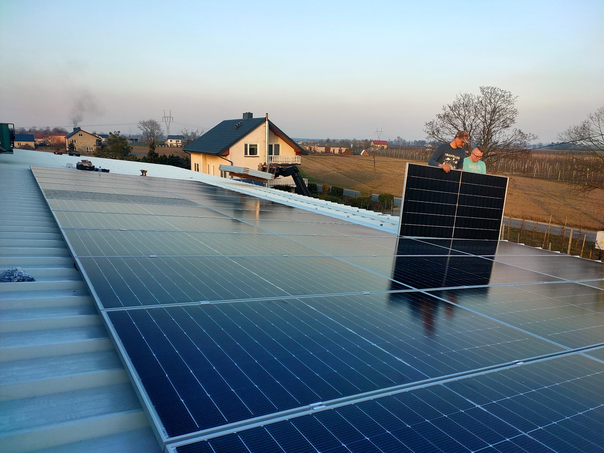 Montaż paneli słonecznych na dachu domu jednorodzinnego; dwóch instalatorów trzyma panel, w tle zabudowania wiejskie.