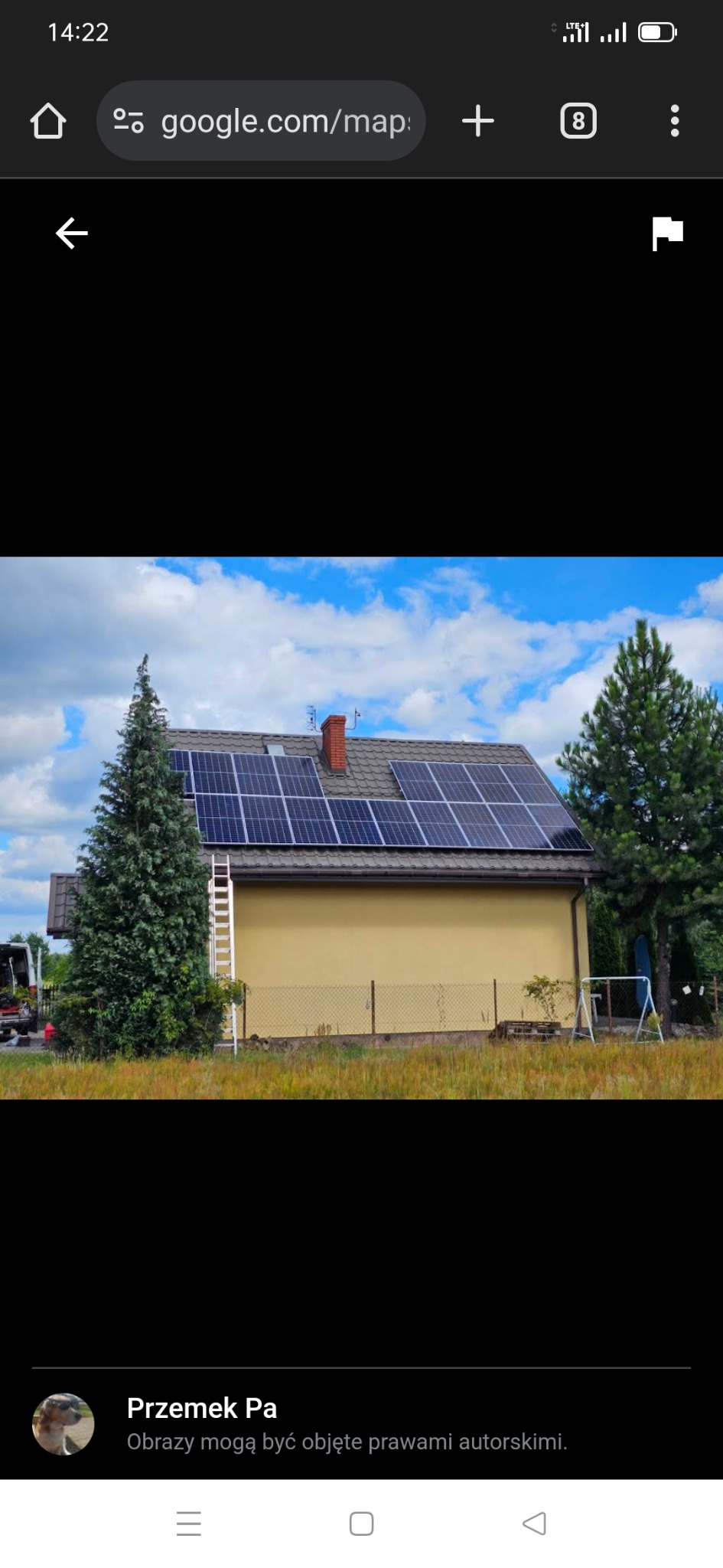 Dom z panelami fotowoltaicznymi na dachu, drabina oparta o ścianę, widok z poziomu gruntu, częściowo zasłonięty przez drzewa iglaste.