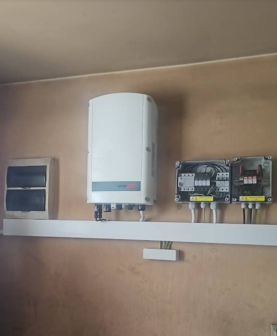 Biały inwerter fotowoltaiczny marki SolarEdge zamontowany na ścianie obok skrzynki elektrycznej i dwóch rozdzielnic w szarych obudowach.