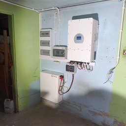 SUNPOWER Sylwester Lemański - Widok na ścianę w garażu lub piwnicy z zamontowanym inwerterem, akumulatorem Power Lab, rozdzielnicami elektrycznymi i przewodami. Ściana pomalowana na niebiesko i zielono, widoczne ślady...
