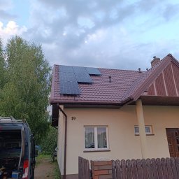 SUNPOWER Sylwester Lemański - Panele słoneczne zamontowane na brązowym dachu domu jednorodzinnego z numerem 19, na tle pochmurnego nieba i drzew, z widocznym samochodem dostawczym z bagażnikiem dachowym.