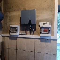 SUNPOWER Sylwester Lemański - Szary falownik firmy Solplanet zamontowany na szorstkiej, beżowej ścianie obok dwóch skrzynek z bezpiecznikami. Widoczne przewody, listwa i fragment rury.