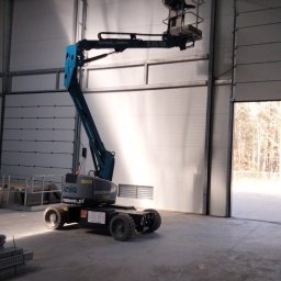 SUNPOWER Sylwester Lemański - Niebieski podnośnik koszowy Genie Z-33/18 wewnątrz hali z segmentową bramą garażową, widoczne elementy konstrukcji dachu i profile stalowe na posadzce.