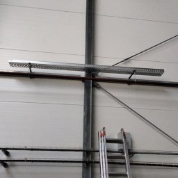 SUNPOWER Sylwester Lemański - Wnętrze hali przemysłowej z widocznymi elementami instalacji: rury, korytka kablowe zawieszone pod sufitem, stalowe liny i drabina oparta o słup konstrukcyjny.