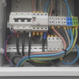 SUNPOWER Sylwester Lemański - Wnętrze skrzynki elektrycznej z bezpiecznikami, różnokolorowymi przewodami i zabezpieczeniami przepięciowymi. Widoczna tabliczka znamionowa SPZ - Tablica przełącznika agregat/sieć.