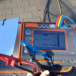 Miernik rezystancji izolacji MPI-540-PV z podłączonymi przewodami pomiarowymi, wskazujący wynik powyżej 2,00 GΩ, leżący na szarym chodniku obok notatnika z logotypem firmy.