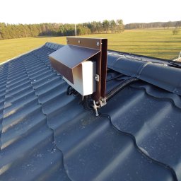 SUNPOWER Sylwester Lemański - Szczegółowe ujęcie elementu instalacji na grafitowym dachu z blachodachówki z widocznymi przewodami w peszlu, na tle zielonego pola i lasu.