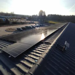 Panele fotowoltaiczne zamontowane na dachu budynku z dachówką ceramiczną, w tle parking z samochodami i stacja benzynowa w słoneczny dzień.