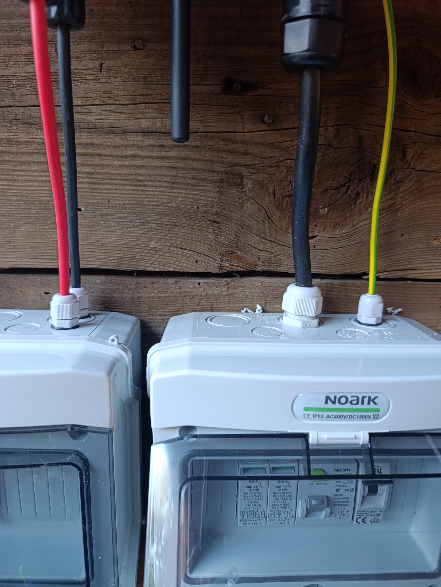 Dwa moduły zabezpieczeń elektrycznych NOARK zamontowane na drewnianej ścianie, z widocznymi przewodami: czerwonym, czarnym i żółto-zielonym.