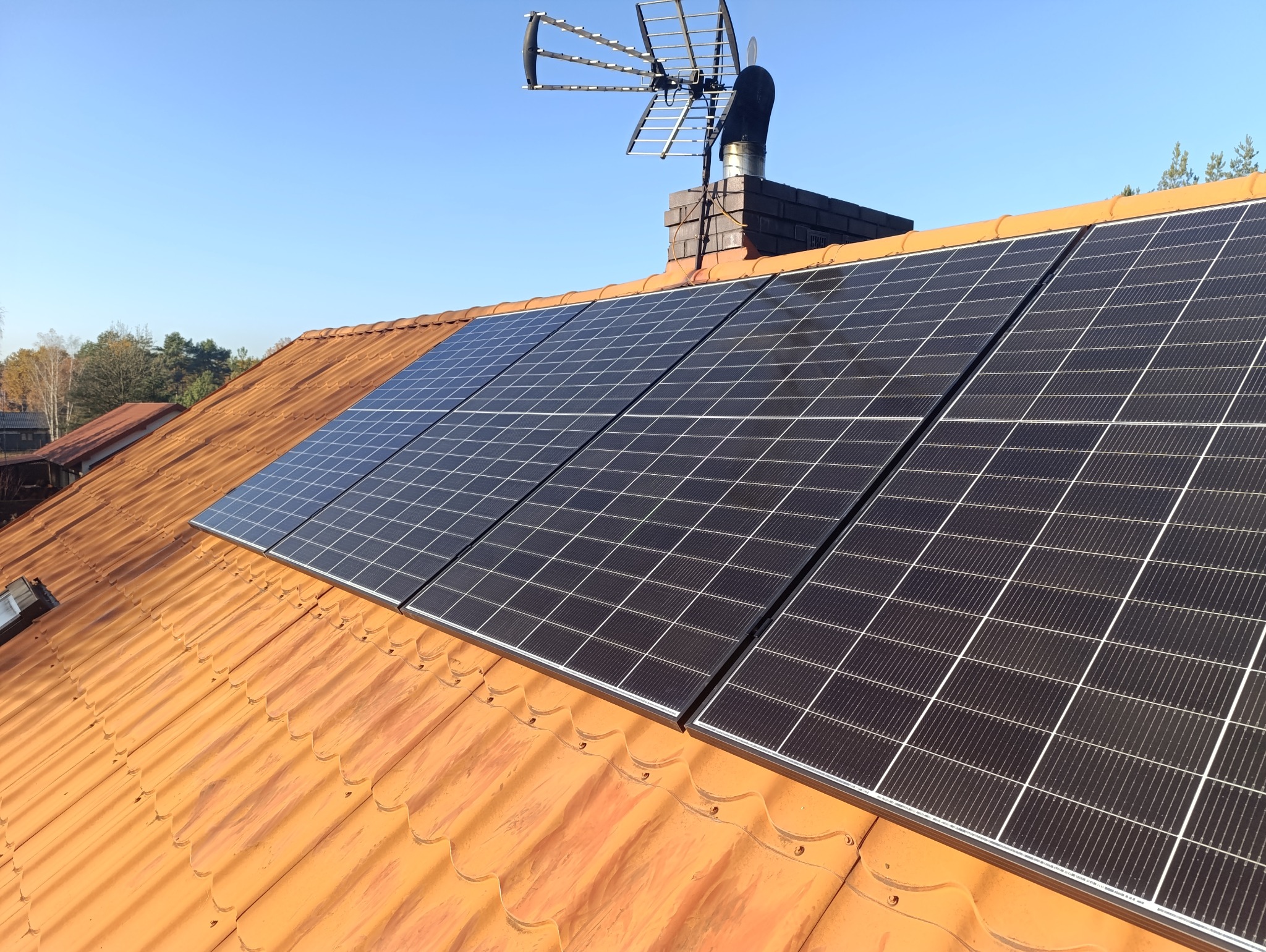 Panele słoneczne zamontowane na pomarańczowym dachu z dachówki ceramicznej, widoczny komin z anteną telewizyjną na tle błękitnego nieba.