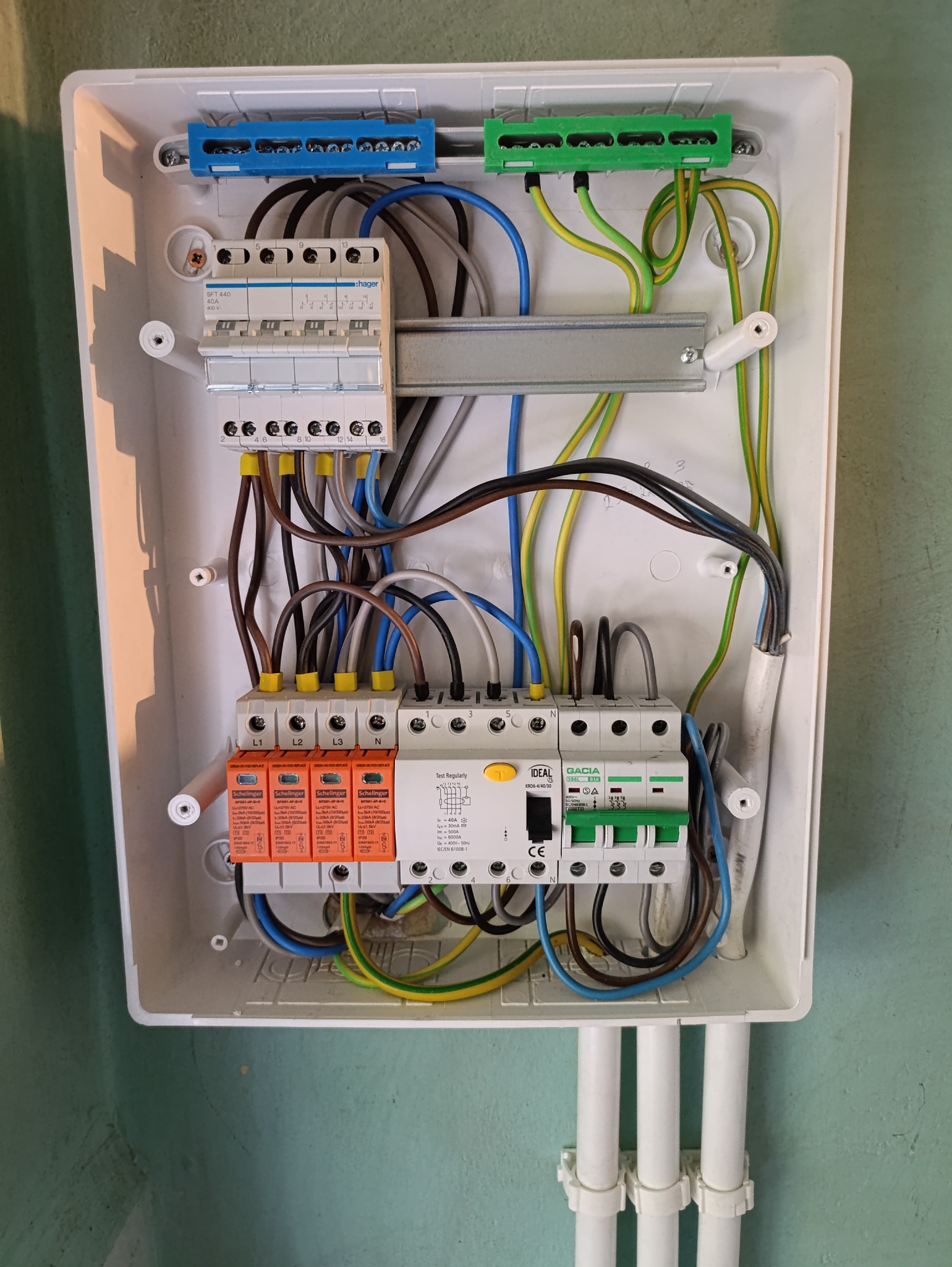 Otwarta skrzynka elektryczna z zainstalowanymi zabezpieczeniami przepięciowymi Schelinger, wyłącznikami Hager i Ideal, widoczne kable w różnych kolorach podłączone do listew zaciskowych, instalacja...