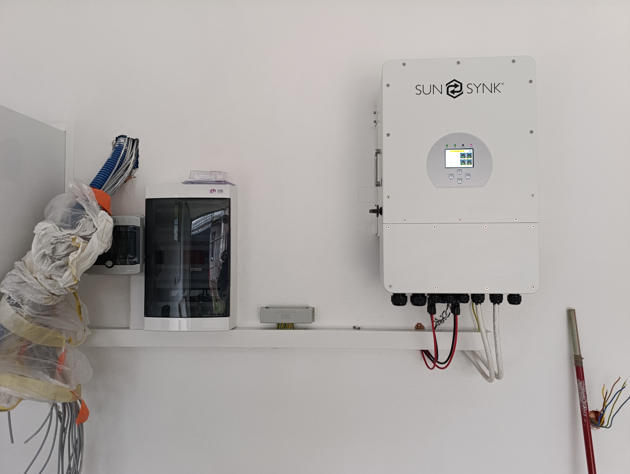 Biały inwerter solarny marki Sunsynk zamontowany na ścianie obok skrzynki elektrycznej i przewodów zabezpieczonych folią. Widoczne kable podłączeniowe i narzędzie.