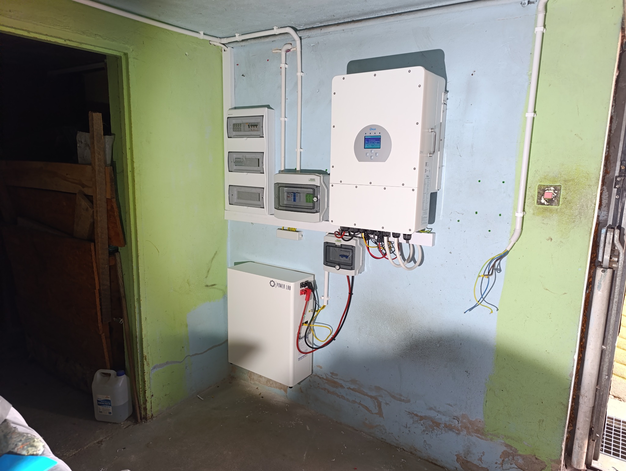 Widok na ścianę w garażu lub piwnicy z zamontowanym inwerterem, akumulatorem Power Lab, rozdzielnicami elektrycznymi i przewodami. Ściana pomalowana na niebiesko i zielono, widoczne ślady...