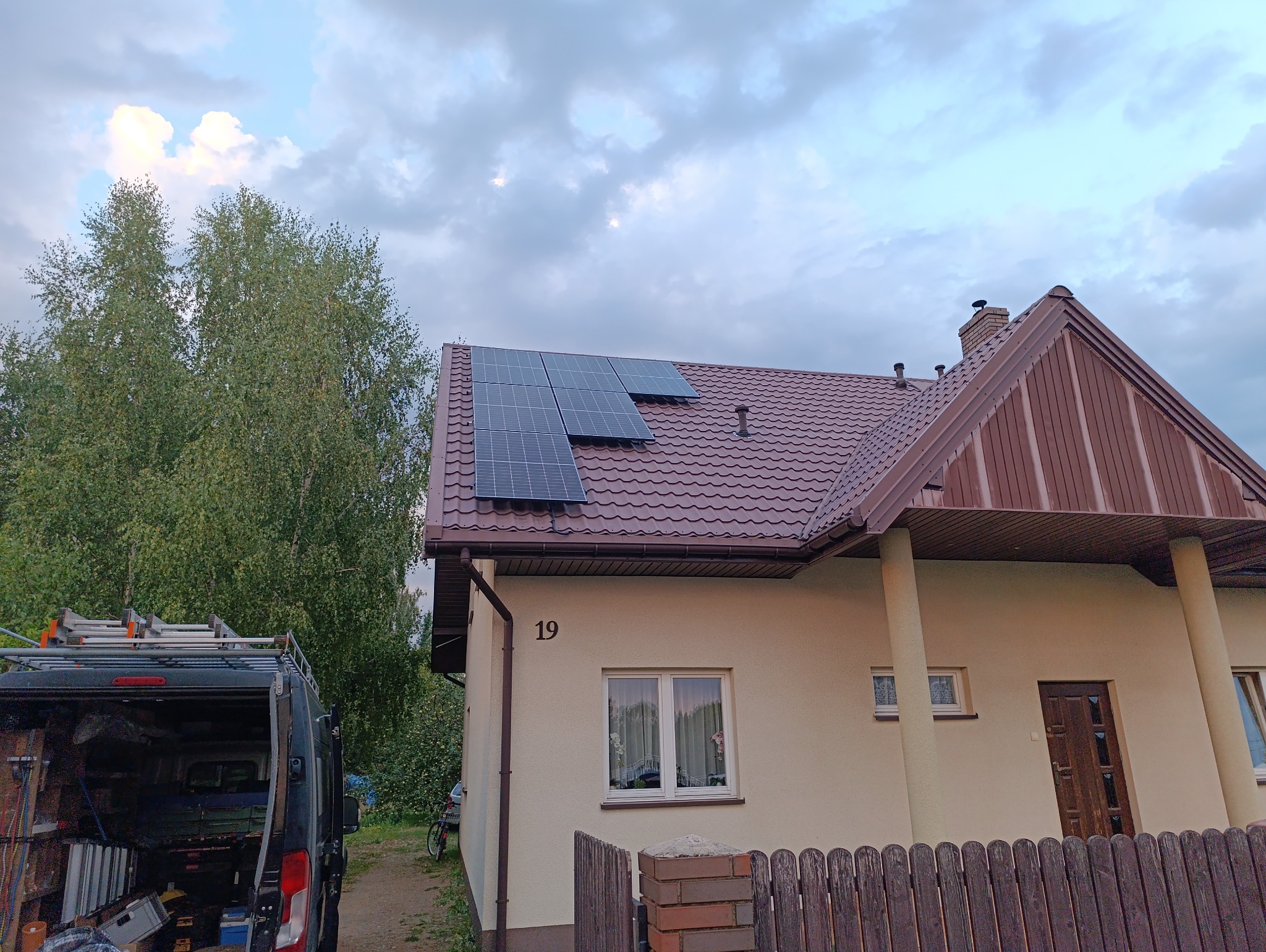 Panele słoneczne zamontowane na brązowym dachu domu jednorodzinnego z numerem 19, na tle pochmurnego nieba i drzew, z widocznym samochodem dostawczym z bagażnikiem dachowym.