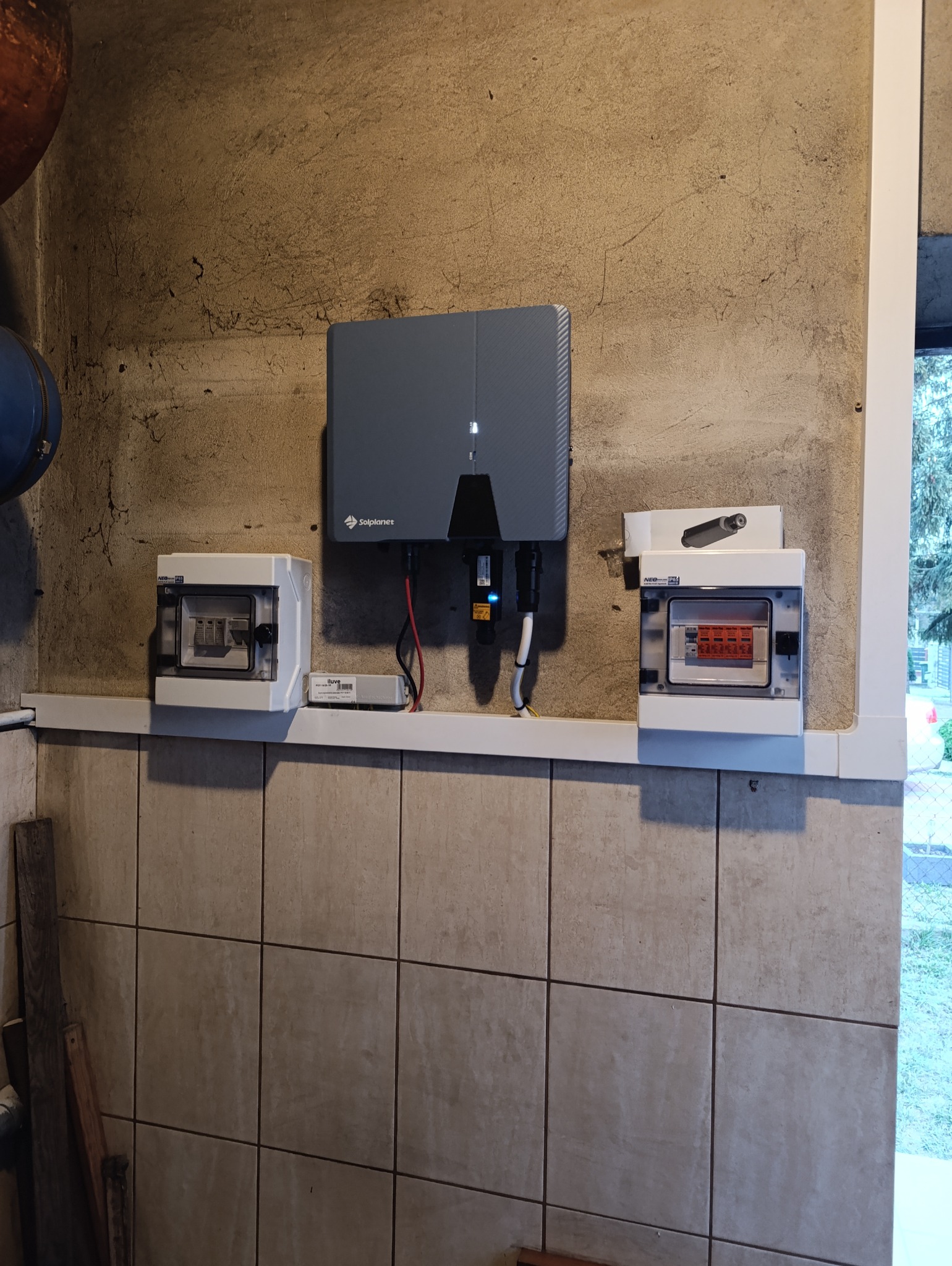 Szary falownik firmy Solplanet zamontowany na szorstkiej, beżowej ścianie obok dwóch skrzynek z bezpiecznikami. Widoczne przewody, listwa i fragment rury.