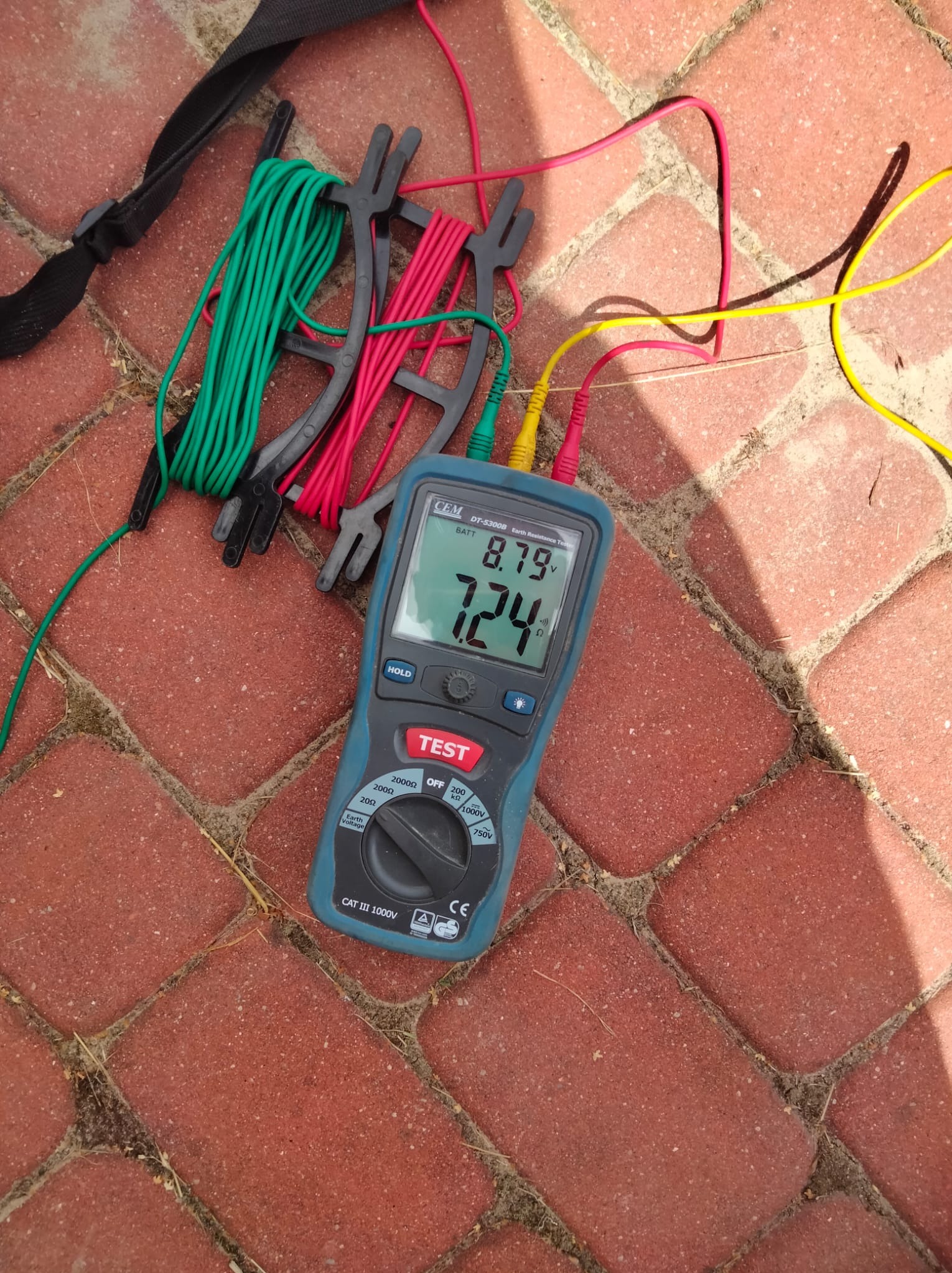 Miernik rezystancji uziomu CEM DT-5300B leżący na czerwonej kostce brukowej, z podłączonymi przewodami pomiarowymi w kolorach zielonym, żółtym i czerwonym oraz zwiniętymi przewodami na uchwytach.