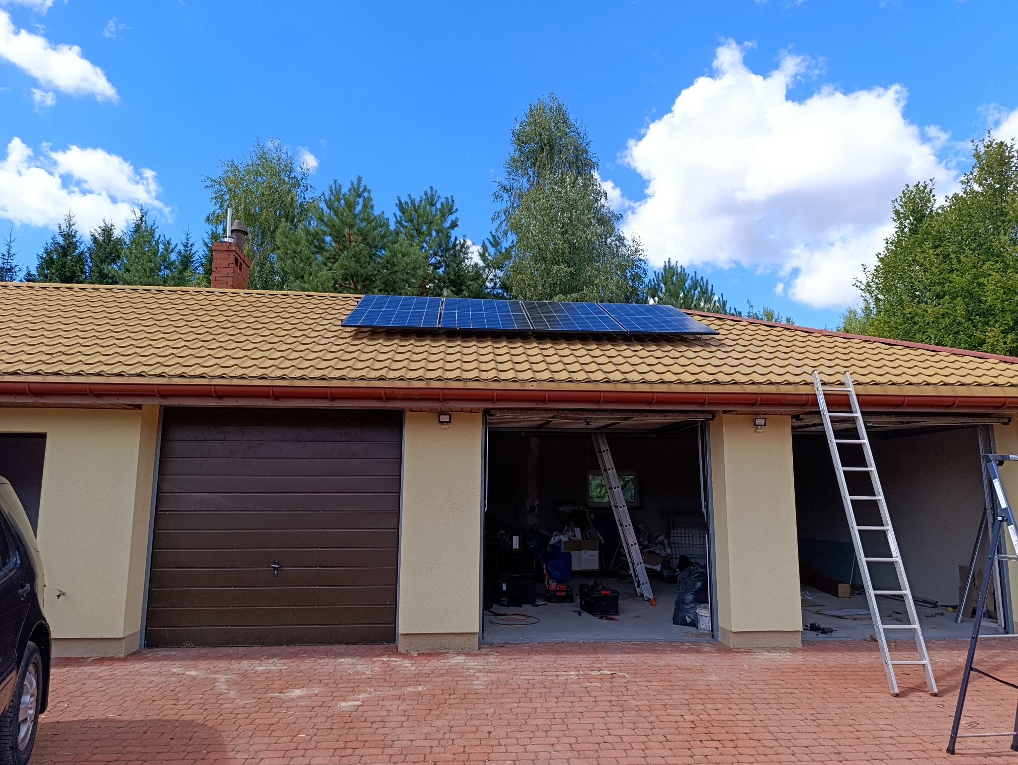 Panele słoneczne zainstalowane na dachu garażu z brązową dachówką, drabina oparta o budynek, widoczne narzędzia w otwartym garażu, błękitne niebo z chmurami w tle.