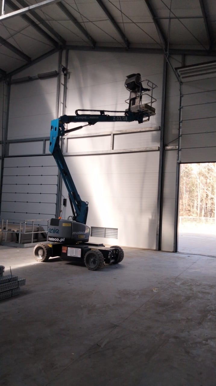 Niebieski podnośnik koszowy Genie Z-33/18 wewnątrz hali z segmentową bramą garażową, widoczne elementy konstrukcji dachu i profile stalowe na posadzce.