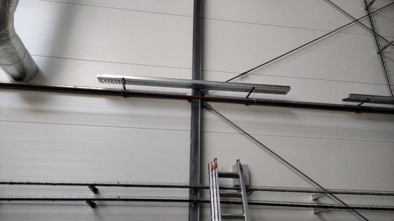 Wnętrze hali przemysłowej z widocznymi elementami instalacji: rury, korytka kablowe zawieszone pod sufitem, stalowe liny i drabina oparta o słup konstrukcyjny.