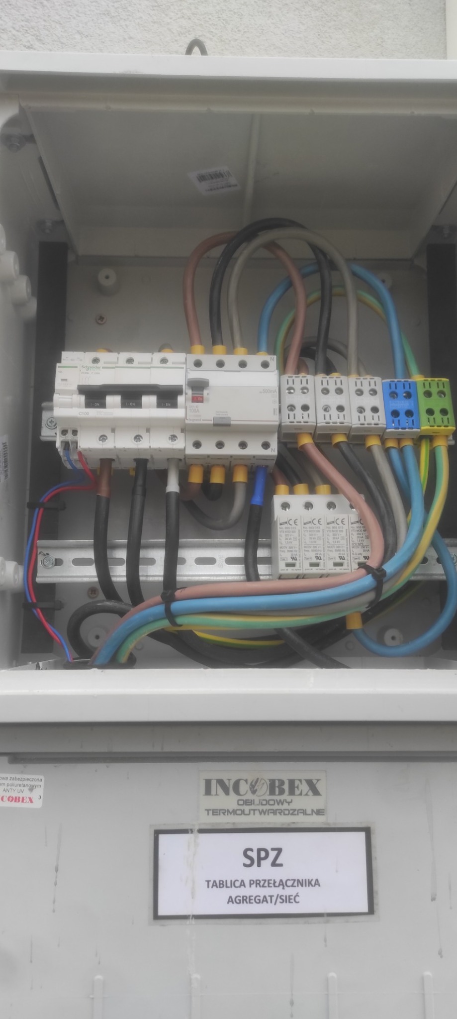 Wnętrze skrzynki elektrycznej z bezpiecznikami, różnokolorowymi przewodami i zabezpieczeniami przepięciowymi. Widoczna tabliczka znamionowa SPZ - Tablica przełącznika agregat/sieć.