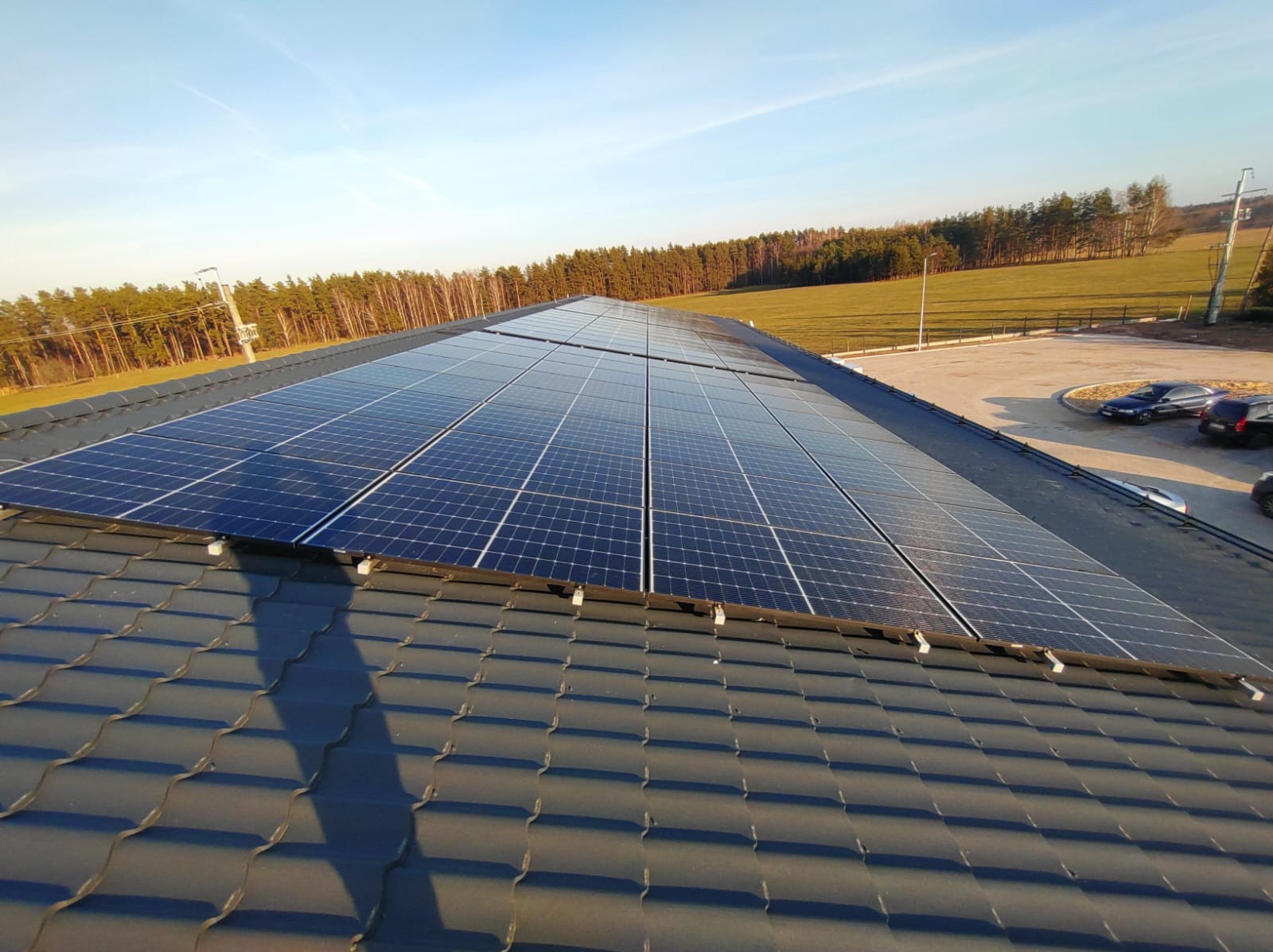 Instalacja paneli słonecznych na dachu pokrytym szarą dachówką, z widokiem na parking, drzewa i błękitne niebo.