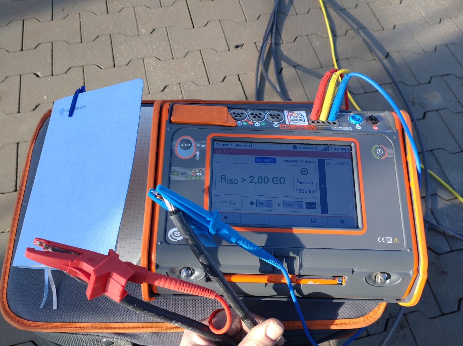 Miernik rezystancji izolacji MPI-540-PV z podłączonymi przewodami pomiarowymi, wskazujący wynik powyżej 2,00 GΩ, leżący na szarym chodniku obok notatnika z logotypem firmy.
