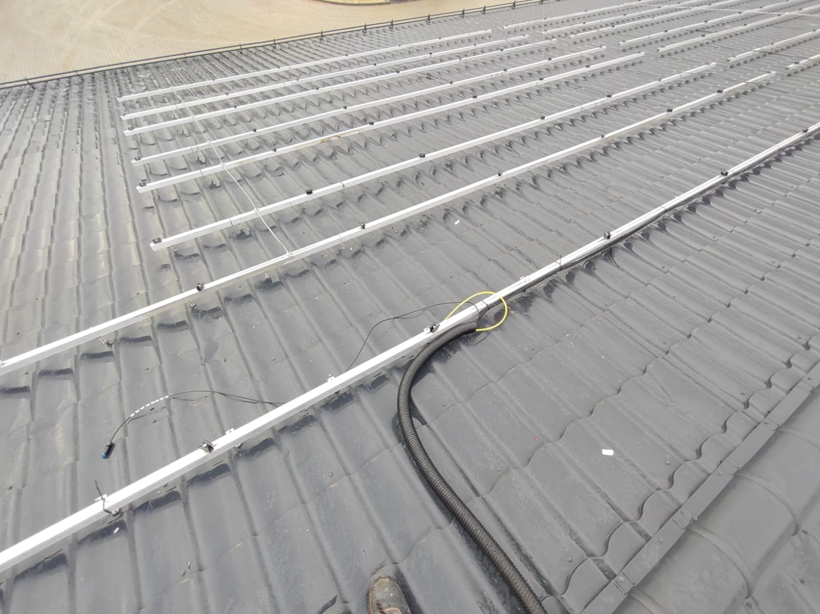 Montaż konstrukcji wsporczej pod panele słoneczne na dachu pokrytym dachówką ceramiczną; widoczne aluminiowe profile mocujące, kable elektryczne i peszel ochronny.