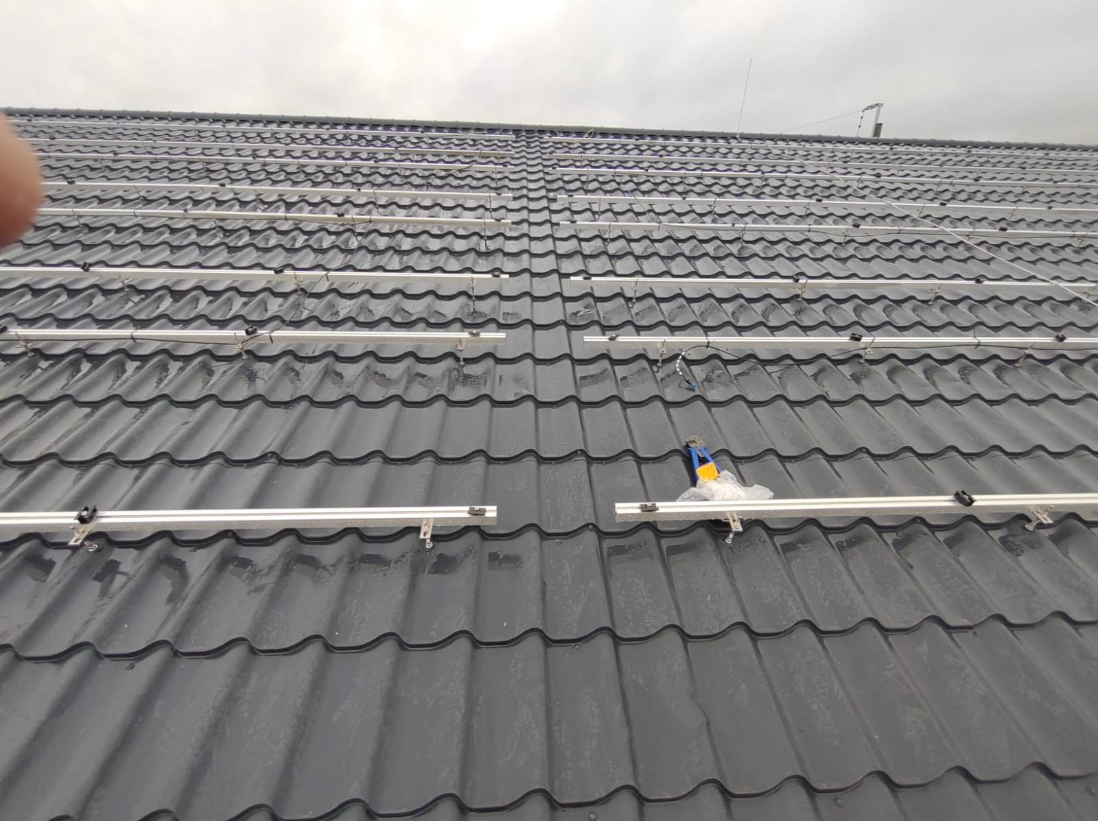 Montaż szyn pod panele fotowoltaiczne na mokrym, grafitowym dachu z dachówki ceramicznej, widoczne narzędzia i przewody.