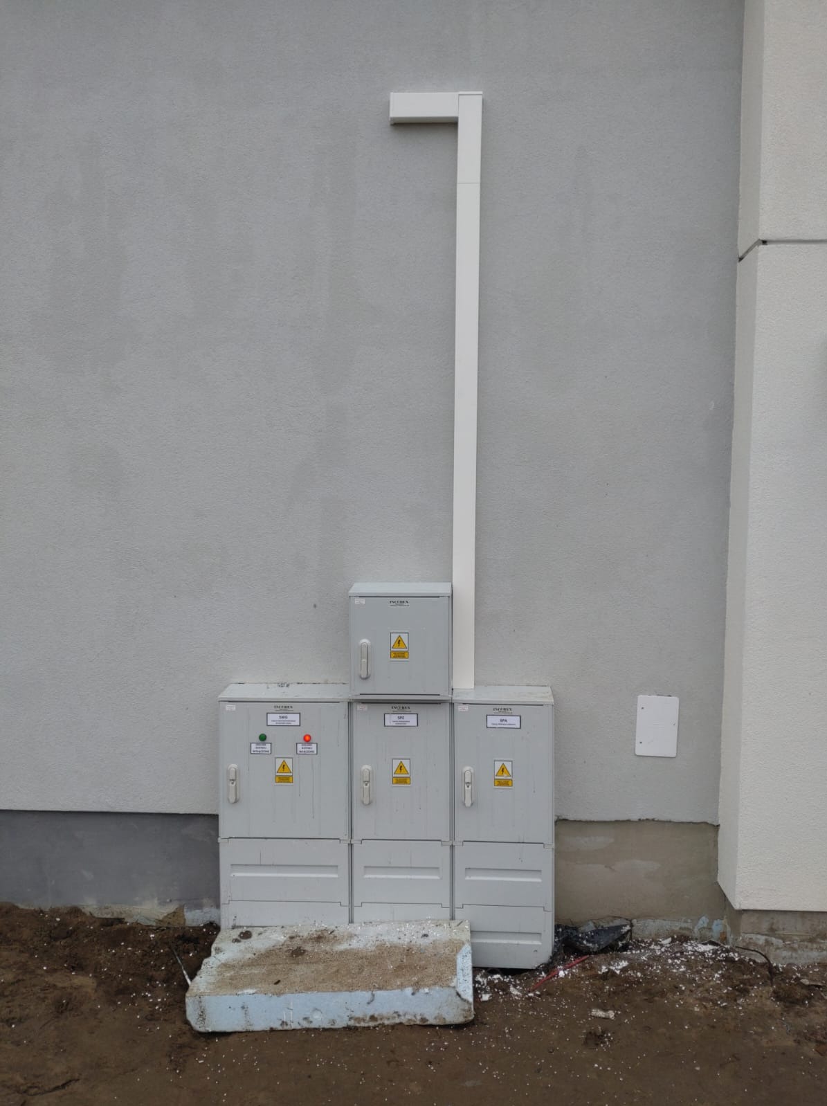 Zestaw szarych skrzynek elektrycznych Insebox z ostrzeżeniami o napięciu, umieszczonych na zewnątrz budynku z szarą elewacją, połączonych białą rurą PCV. Widoczne okablowanie i fragment fundamentu.