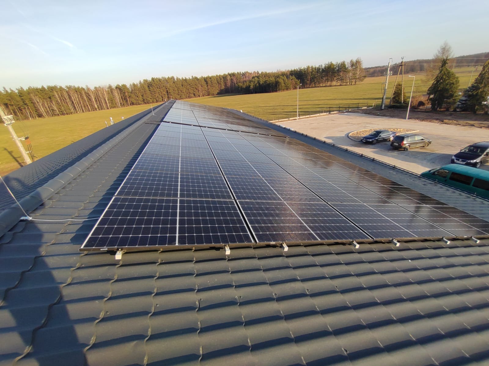 Panele słoneczne zamontowane na ciemnoszarym dachu z dachówki bitumicznej, w tle zielone pola i drzewa, a także parking z samochodami w słoneczny dzień.