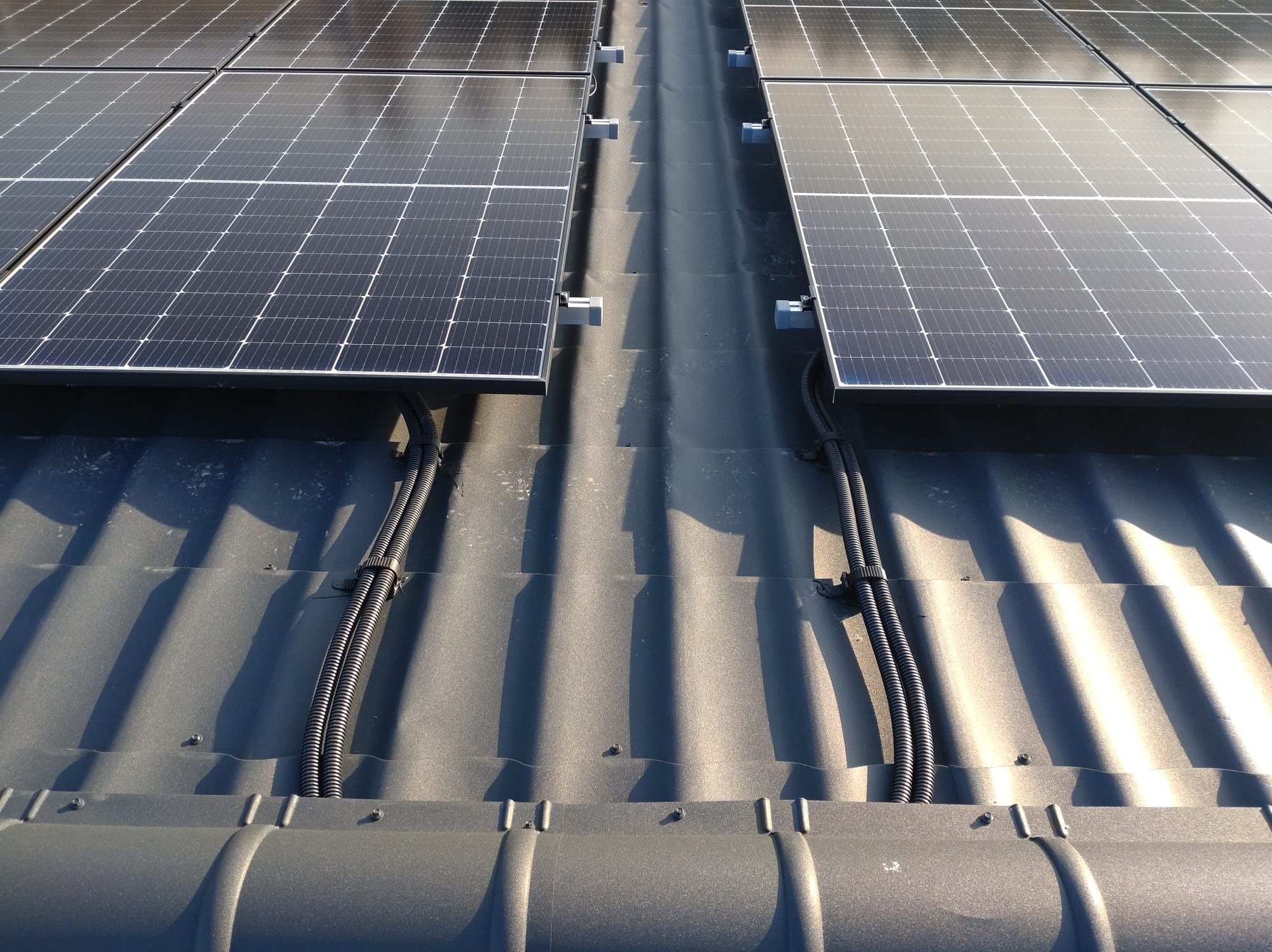 Panele słoneczne zamontowane na metalowym dachu, widoczne czarne rury ochronne prowadzące kable elektryczne, przymocowane do dachu uchwytami.