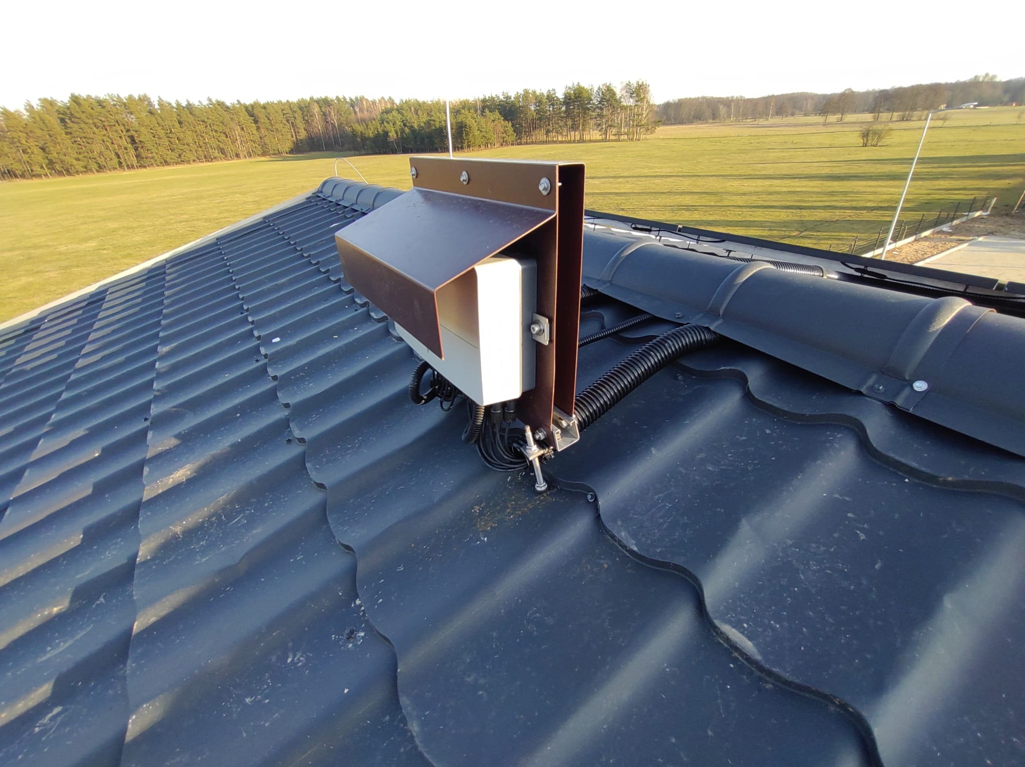 Szczegółowe ujęcie elementu instalacji na grafitowym dachu z blachodachówki z widocznymi przewodami w peszlu, na tle zielonego pola i lasu.