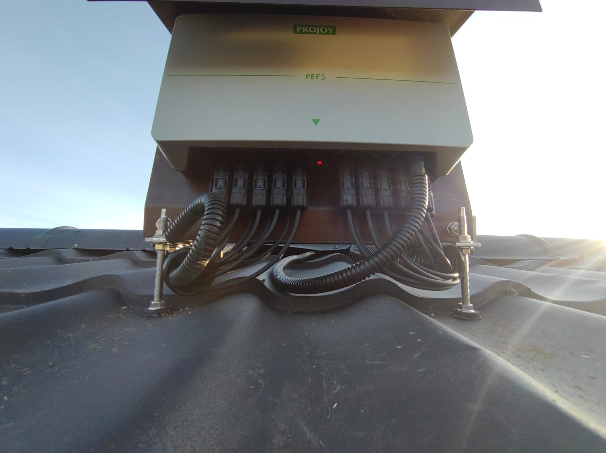 Szczegółowe ujęcie inwertera marki Projoy PEFS zamontowanego na czarnym dachu z falistej blachy, z widocznymi przewodami w peszlach i elementami mocującymi.