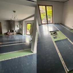 Układanie folii grzewczej HEAT-ON na podłodze w przestronnym pokoju z kominkiem i dużym oknem. Prace w toku, widoczny instalator. Etap przygotowania do dalszych prac wykończeniowych.