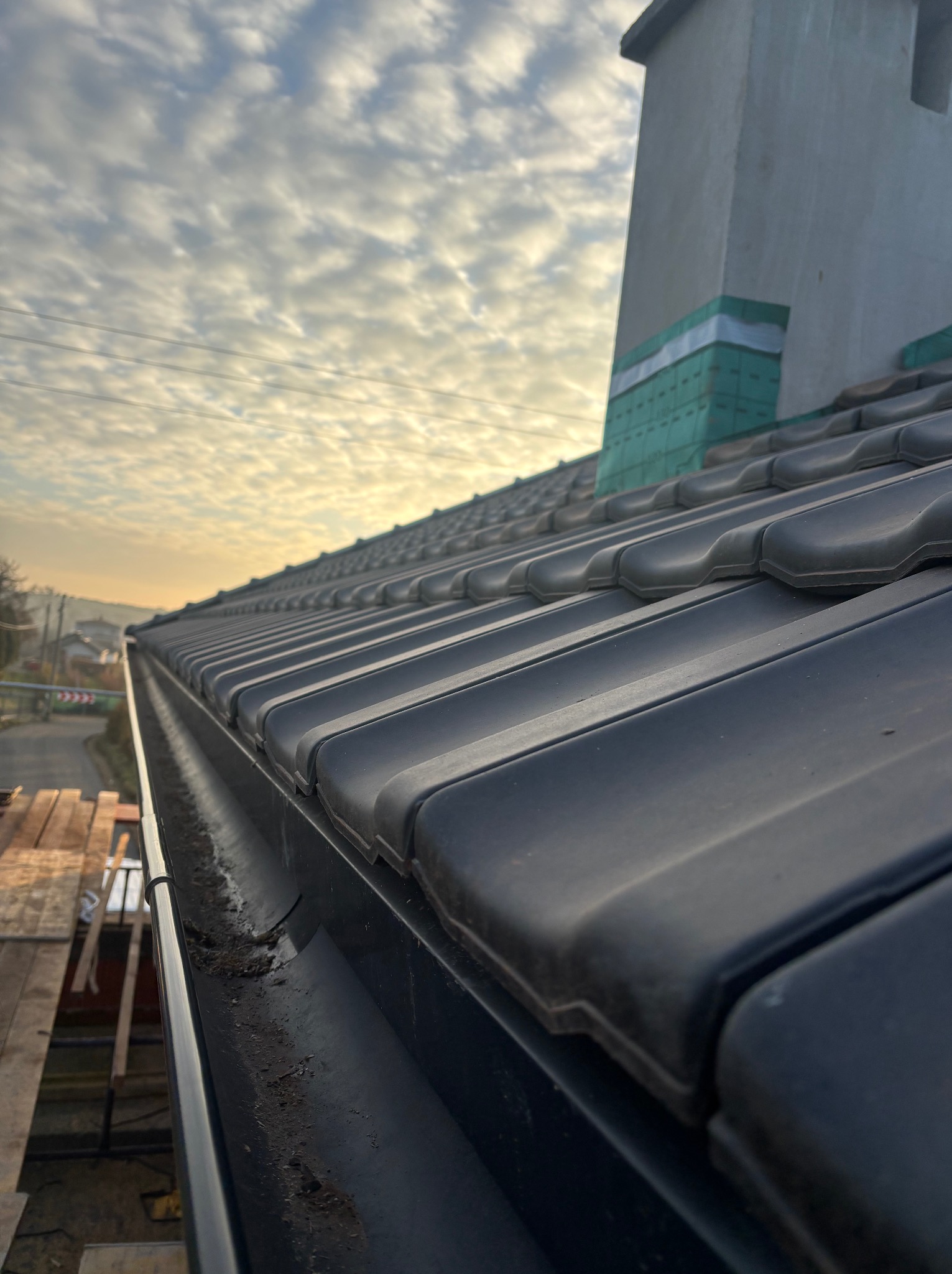 Czarny dach z ceramiczną dachówką i rynną, widok z dołu na tle pochmurnego nieba. Widoczne elementy komina i rusztowania. Ujęcie z bliska.