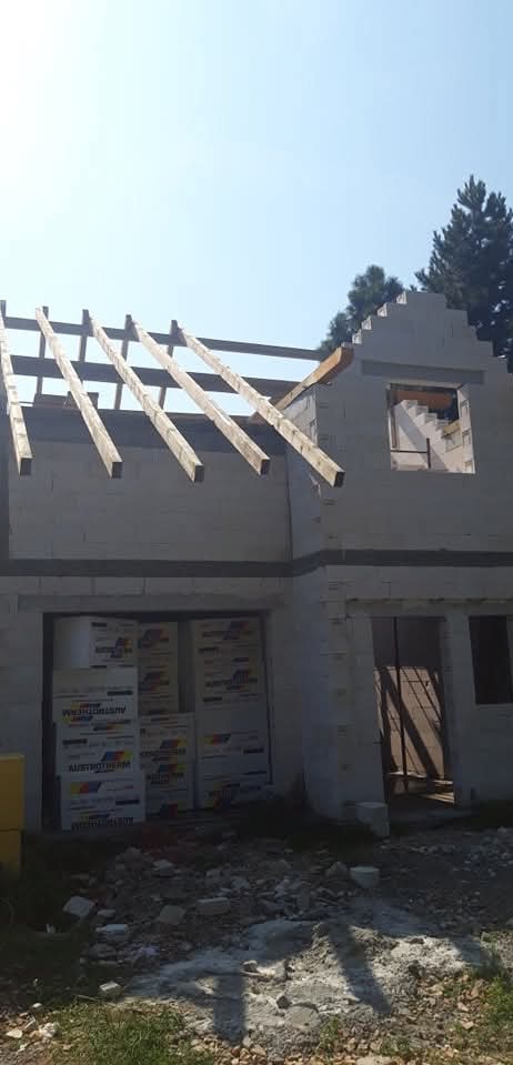 Budowa domu w stanie surowym otwartym: ściany z bloczków, drewniana konstrukcja dachu, okna bez szyb, materiały izolacyjne na posesji. Słoneczny dzień.